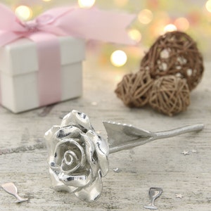 Op de afbeelding: Een zilveren roos met een steel en bladeren. De roos ligt op een wit houten oppervlak met een witte geschenkdoos en bruine wilgenbollen op de achtergrond.
