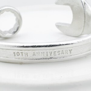 May include: A silver-colored wrench-shaped bracelet. The bracelet has the words "10TH ANNIVERSARY" engraved on the band. The wrench design includes a circular end and an open-ended wrench shape.