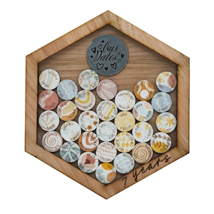 Puede incluir: Un marco hexagonal de madera con un frente de vidrio muestra una colección de discos redondos, coloridos y pintados con fechas y diseños escritos a mano. El marco tiene un círculo de metal en el centro con las palabras "Our Dates" y un número "7" grabado debajo del marco, que indica "7 años".