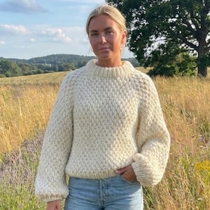 May include: A cream-coloured, textured knit sweater with a high neckline and long sleeves. The sweater is paired with light blue jeans and black wellington boots. The background features a field of tall grass and a large tree under a blue sky.