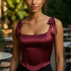 May include: A sleeveless burgundy satin corset top featuring a square neckline and shoulder straps with decorative tie details. The bodice has a structured, fitted silhouette with a pointed hemline that tapers at the waist, crafted from a smooth fabric.