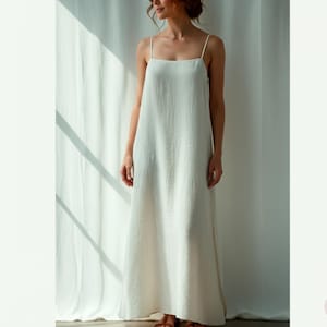 Puede incluir: Un vestido largo color blanco roto con finos tirantes. El vestido tiene un diseño sencillo y fluido y un escote cuadrado. La tela parece ser de lino o algodón ligero, con una textura sutil. El vestido lo lleva una persona de pie frente a una cortina blanca.