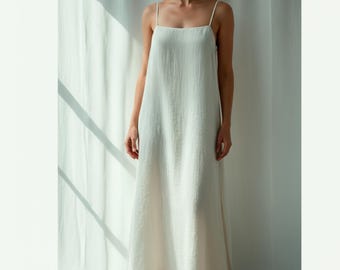 May include: A floor-length, off-white dress with thin spaghetti straps. The dress has a simple, flowing design and a square neckline. The fabric appears to be a lightweight linen or cotton, with a subtle texture. The dress is worn by a person standing in front of a white curtain.