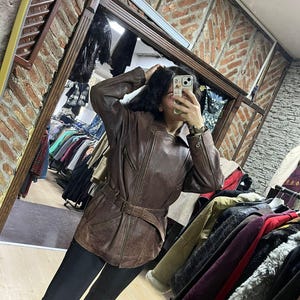 May include: A brown leather jacket with a belted waist and zipper details. The jacket has a classic design and appears to be vintage. The image is taken in a clothing store, with racks of clothing visible in the background.