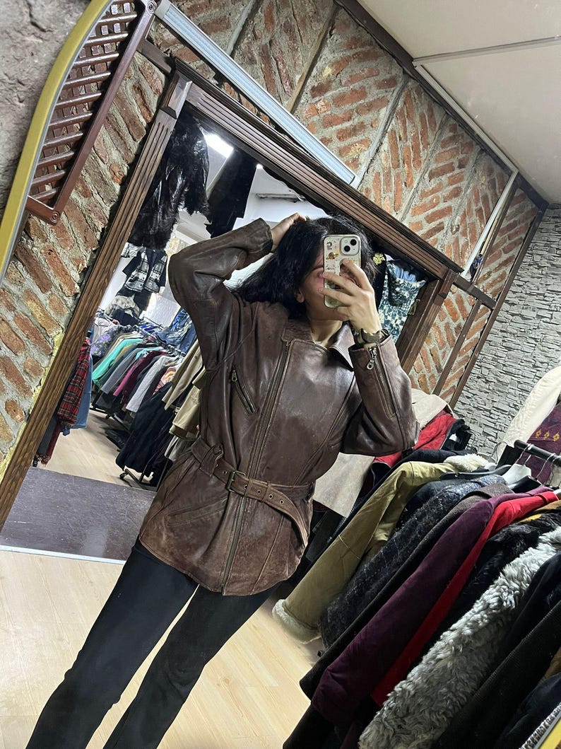 May include: A brown leather jacket with a belted waist and zipper details. The jacket has a classic design and appears to be vintage. The image is taken in a clothing store, with racks of clothing visible in the background.