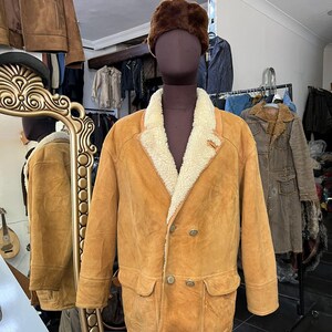 May include: A tan suede jacket with a cream shearling lining and a brown fur hat. The jacket has a double-breasted design with two rows of buttons and two front pockets. The jacket is displayed on a mannequin in a shop setting.