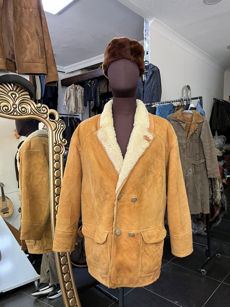 May include: A tan suede jacket with a cream shearling lining and a brown fur hat. The jacket has a double-breasted design with two rows of buttons and two front pockets. The jacket is displayed on a mannequin in a shop setting.