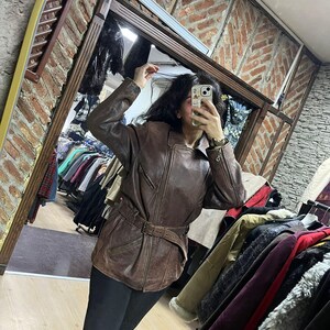 May include: Brown leather jacket with a belted waist and zipper details. The jacket has a classic design and a vintage look. The image is taken in a clothing store, with racks of clothing visible in the background.
