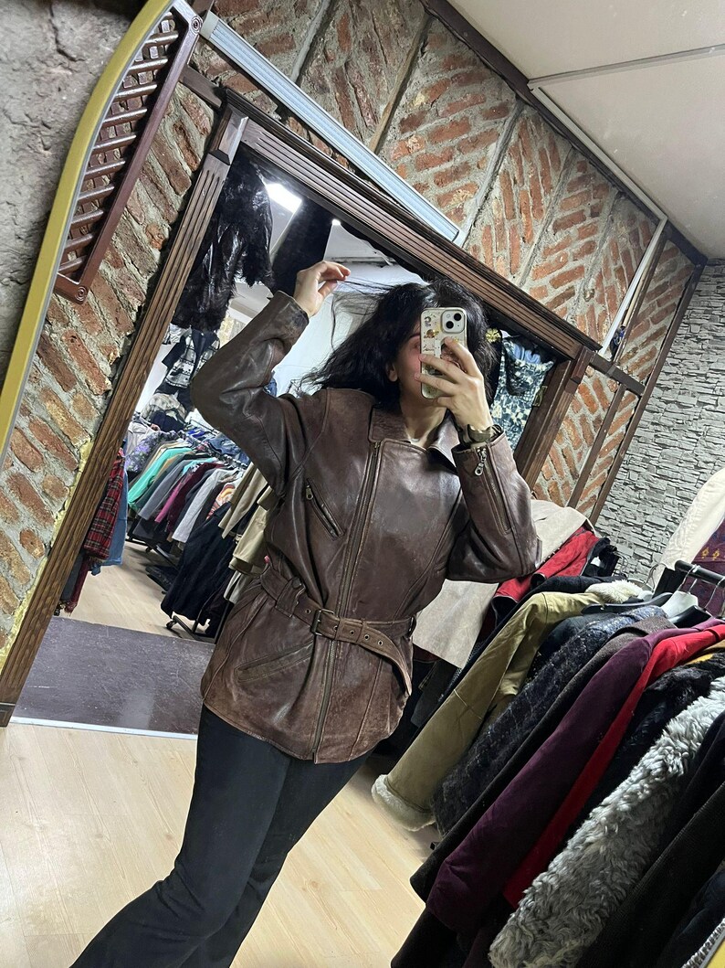 May include: Brown leather jacket with a belted waist and zipper details. The jacket has a classic design and a vintage look. The image is taken in a clothing store, with racks of clothing visible in the background.