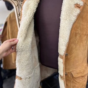 May include: A tan suede jacket with a cream shearling lining. The jacket has a button closure, two front pockets, and a classic collar. The interior lining is visible, offering warmth and comfort.