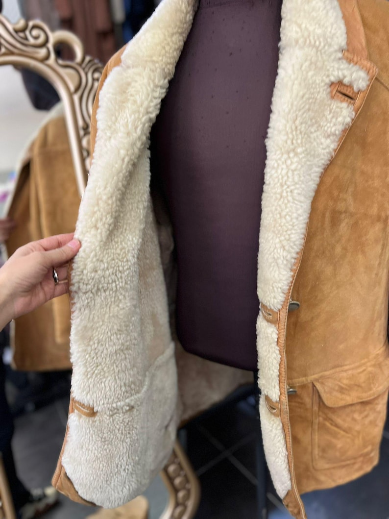 May include: A tan suede jacket with a cream shearling lining. The jacket has a button closure, two front pockets, and a classic collar. The interior lining is visible, offering warmth and comfort.