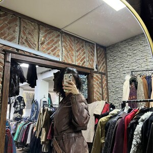 May include: A brown leather jacket with a belted waist and zipper details. The jacket is displayed in a vintage clothing store, with racks of various coats and jackets visible in the background. The mirror reflects the store's interior.