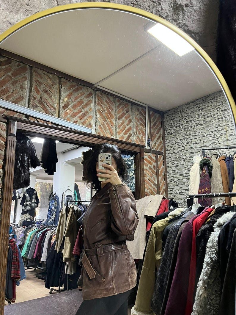 May include: A brown leather jacket with a belted waist and zipper details. The jacket is displayed in a vintage clothing store, with racks of various coats and jackets visible in the background. The mirror reflects the store's interior.