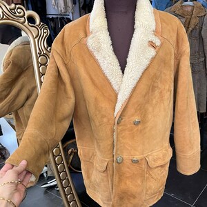May include: A tan suede jacket with a cream shearling collar and lining. The double-breasted jacket has two front pockets and silver buttons. The jacket is displayed on a mannequin.