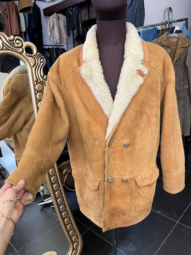 May include: A tan suede jacket with a cream shearling collar and lining. The double-breasted jacket has two front pockets and silver buttons. The jacket is displayed on a mannequin.