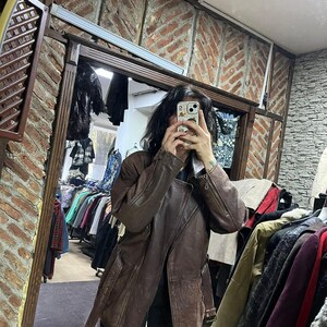 May include: A brown leather jacket with a belted waist and asymmetrical zipper. The jacket has a classic design with a notched collar and long sleeves. The jacket is displayed in a clothing store, with racks of clothing visible in the background.