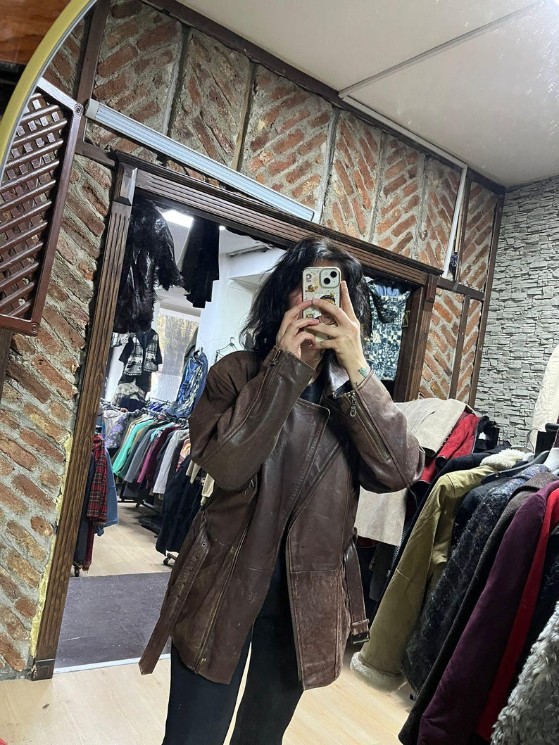 May include: A brown leather jacket with a belted waist and asymmetrical zipper. The jacket has a classic design with a notched collar and long sleeves. The jacket is displayed in a clothing store, with racks of clothing visible in the background.