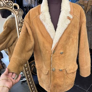 May include: A tan suede jacket with a cream shearling lining and collar. The jacket has a double-breasted design with two rows of buttons, two front pockets, and a small leather tag. The jacket is displayed on a mannequin.