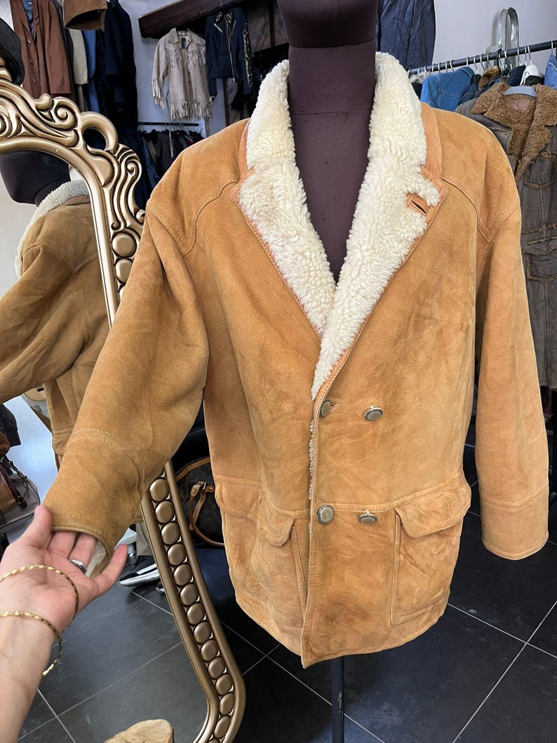 May include: A tan suede jacket with a cream shearling lining and collar. The jacket has a double-breasted design with two rows of buttons, two front pockets, and a small leather tag. The jacket is displayed on a mannequin.