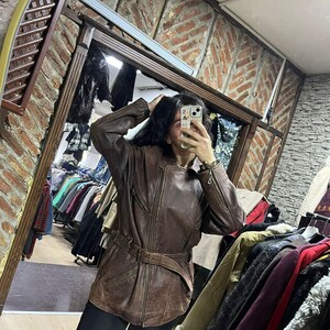 May include: A brown leather jacket with a belted waist and zipper details. The jacket has a vintage look and is worn over black pants. The image is taken in a clothing store, with racks of clothing visible in the background.