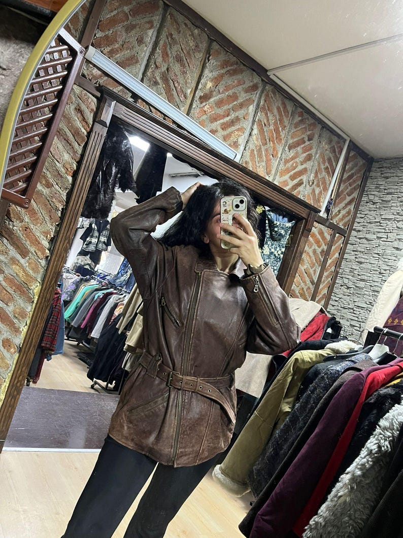 May include: A brown leather jacket with a belted waist and zipper details. The jacket has a vintage look and is worn over black pants. The image is taken in a clothing store, with racks of clothing visible in the background.