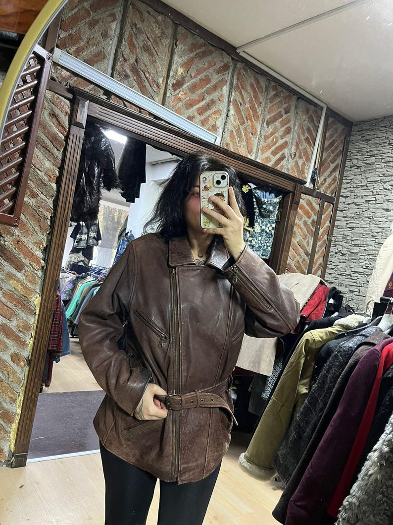 May include: A brown leather jacket with a belted waist and a zipper closure. The jacket has a classic design with a collar and pockets. The leather appears worn, giving it a vintage look. The jacket is being worn in a shop.