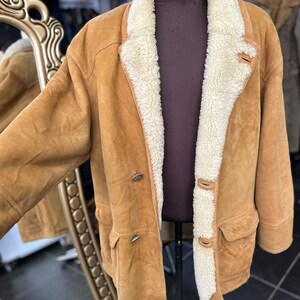 May include: A tan suede jacket with a cream shearling lining and collar. The jacket features two front pockets, two buttons, and a button closure at the collar. The jacket is displayed on a mannequin.
