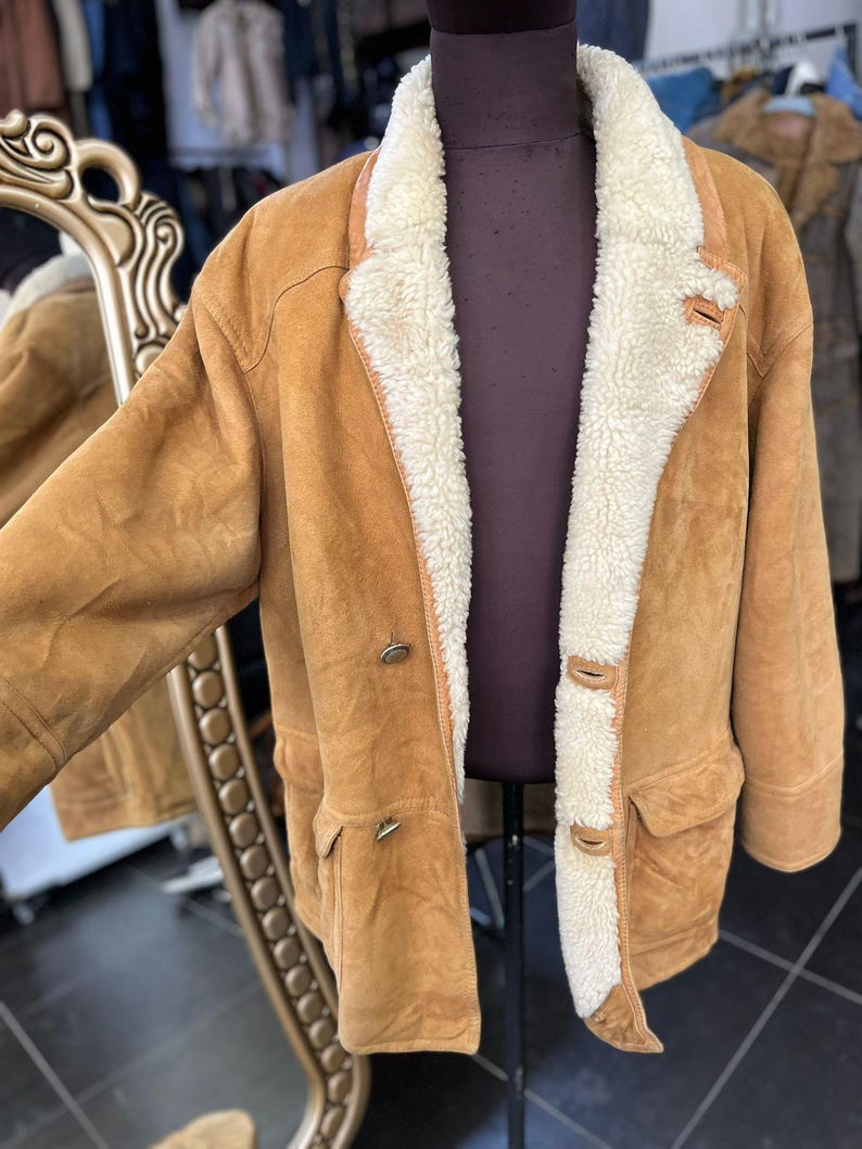 May include: A tan suede jacket with a cream shearling lining and collar. The jacket features two front pockets, two buttons, and a button closure at the collar. The jacket is displayed on a mannequin.
