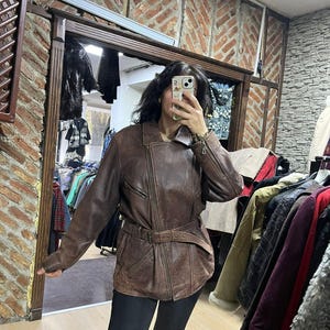 May include: A brown leather jacket with a belted waist and asymmetrical zipper. The jacket has a classic biker style with a notched collar and multiple pockets. The leather appears worn, giving it a vintage look. The jacket is shown in a store setting.