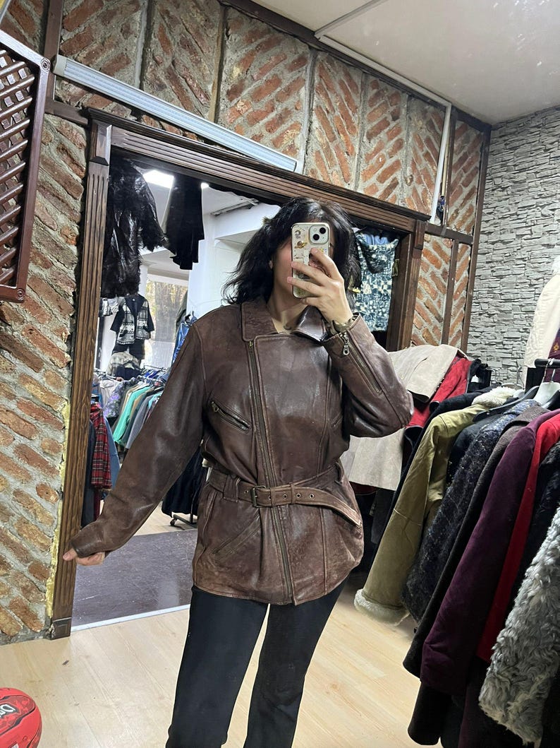May include: A brown leather jacket with a belted waist and asymmetrical zipper. The jacket has a classic biker style with a notched collar and multiple pockets. The leather appears worn, giving it a vintage look. The jacket is shown in a store setting.