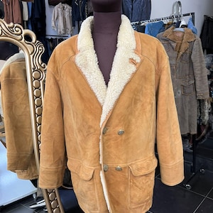 May include: A tan suede coat with a cream shearling lining and collar. The double-breasted coat features two front pockets and silver buttons. The coat is displayed on a mannequin in a clothing store.