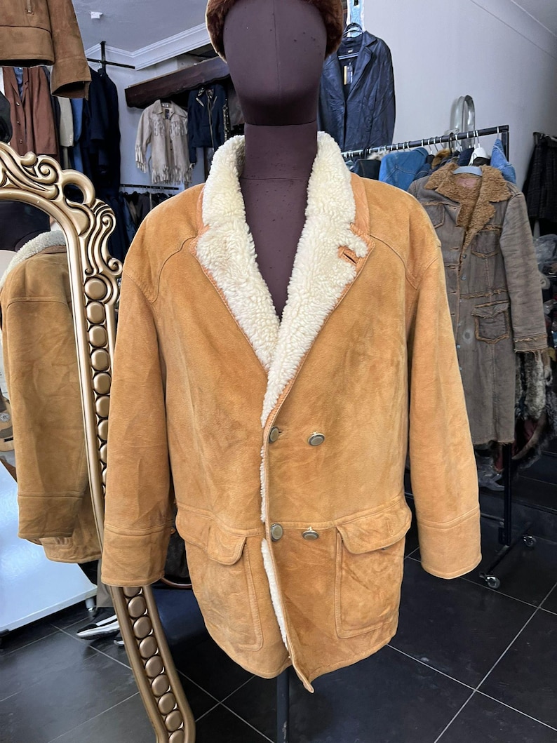 May include: A tan suede coat with a cream shearling lining and collar. The double-breasted coat features two front pockets and silver buttons. The coat is displayed on a mannequin in a clothing store.