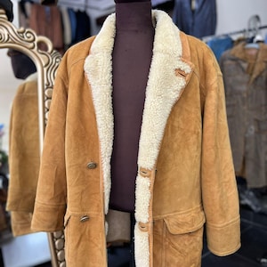 May include: A tan suede coat with a cream shearling lining and collar. The coat has a double-breasted design with two rows of buttons, two front pockets, and a classic, warm aesthetic. The coat is displayed on a mannequin.