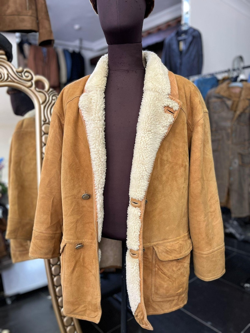 May include: A tan suede coat with a cream shearling lining and collar. The coat has a double-breasted design with two rows of buttons, two front pockets, and a classic, warm aesthetic. The coat is displayed on a mannequin.