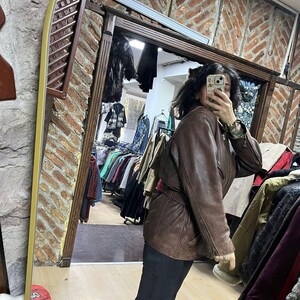 May include: A brown leather jacket with a belted waist. The jacket has a classic design with a collar and pockets. The image is taken in a clothing store, with racks of clothing visible in the background.
