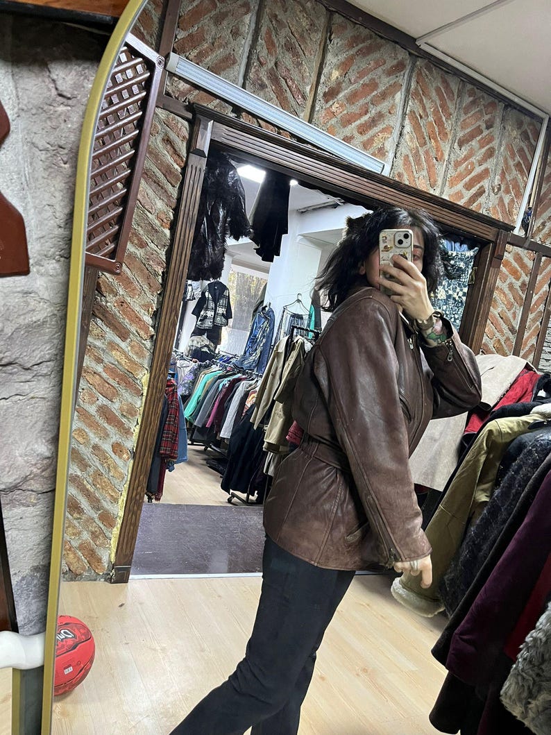 May include: A brown leather jacket with a belted waist. The jacket has a classic design with a collar and pockets. The image is taken in a clothing store, with racks of clothing visible in the background.