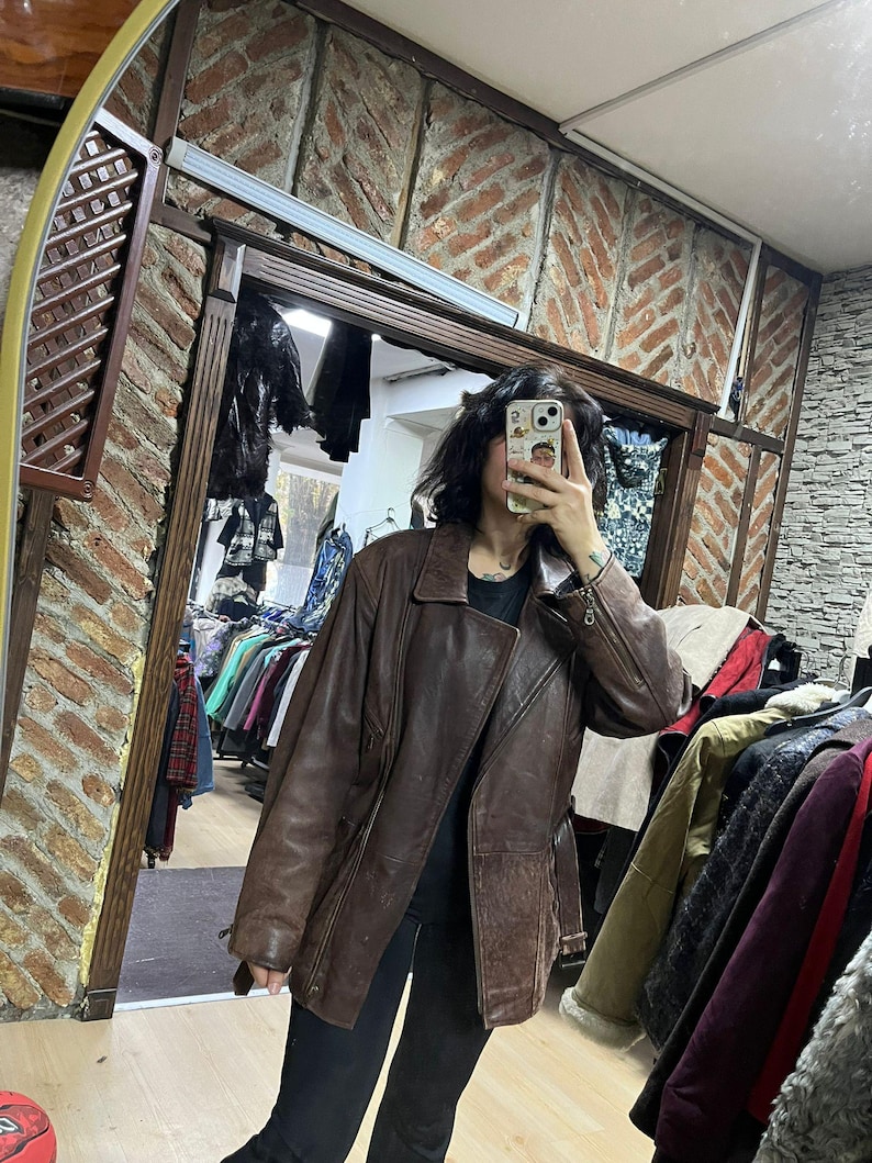 May include: A brown leather jacket with a classic design. The jacket has a notched collar, a button closure, and multiple pockets. The leather appears worn, giving it a vintage look. The jacket is part of a clothing collection.