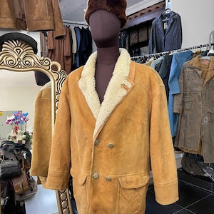May include: A tan suede jacket with a cream shearling collar and lining, displayed on a mannequin. The jacket features two front pockets and a double-breasted button closure. A brown fur hat is on the mannequin's head. Other jackets are visible in the background.