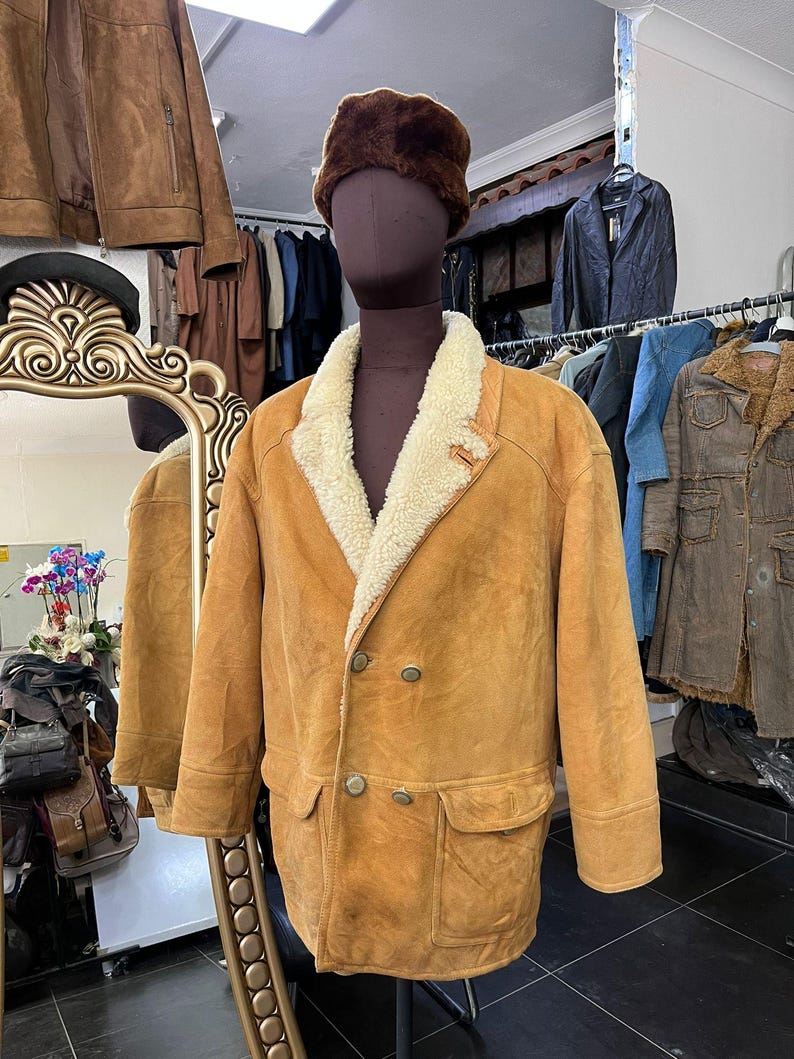 May include: A tan suede jacket with a cream shearling collar and lining, displayed on a mannequin. The jacket features two front pockets and a double-breasted button closure. A brown fur hat is on the mannequin's head. Other jackets are visible in the background.