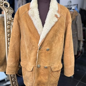 May include: A tan suede jacket with a cream shearling collar and lining. The double-breasted jacket has two front pockets and silver buttons. The jacket is displayed on a mannequin.