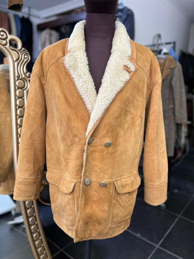 May include: A tan suede jacket with a cream shearling collar and lining. The double-breasted jacket has two front pockets and silver buttons. The jacket is displayed on a mannequin.