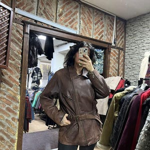 May include: A brown leather jacket with a belted waist and asymmetrical zipper closure. The jacket has a classic biker style with a notched collar and multiple pockets. The leather appears distressed, giving it a vintage look. The jacket is worn over black pants.