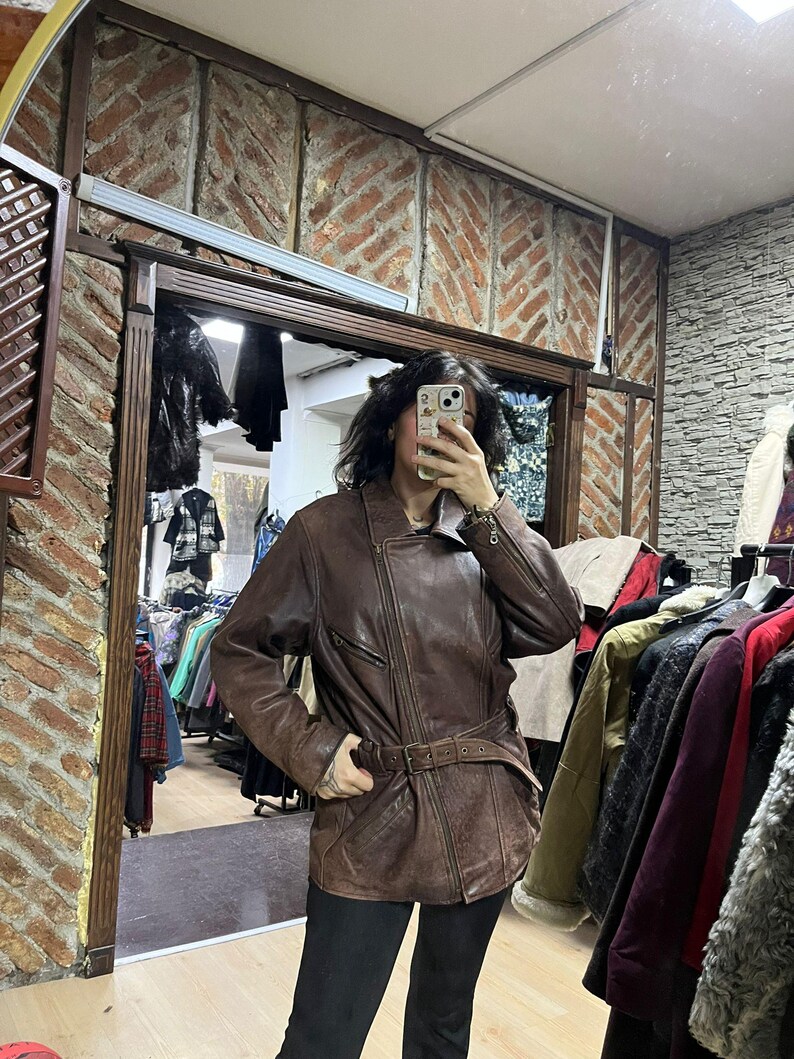 May include: A brown leather jacket with a belted waist and asymmetrical zipper closure. The jacket has a classic biker style with a notched collar and multiple pockets. The leather appears distressed, giving it a vintage look. The jacket is worn over black pants.