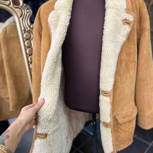 May include: A tan suede coat with a cream shearling lining and trim. The coat has a classic design with a notched collar, button closures, and two front pockets. The coat is displayed on a mannequin.