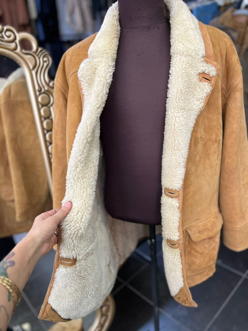 May include: A tan suede coat with a cream shearling lining and trim. The coat has a classic design with a notched collar, button closures, and two front pockets. The coat is displayed on a mannequin.