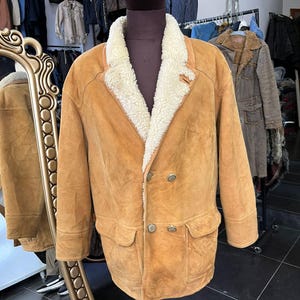 May include: A tan suede jacket with a cream shearling collar and lining. The double-breasted jacket has two front pockets and three buttons. The jacket is displayed on a mannequin in a vintage clothing shop.
