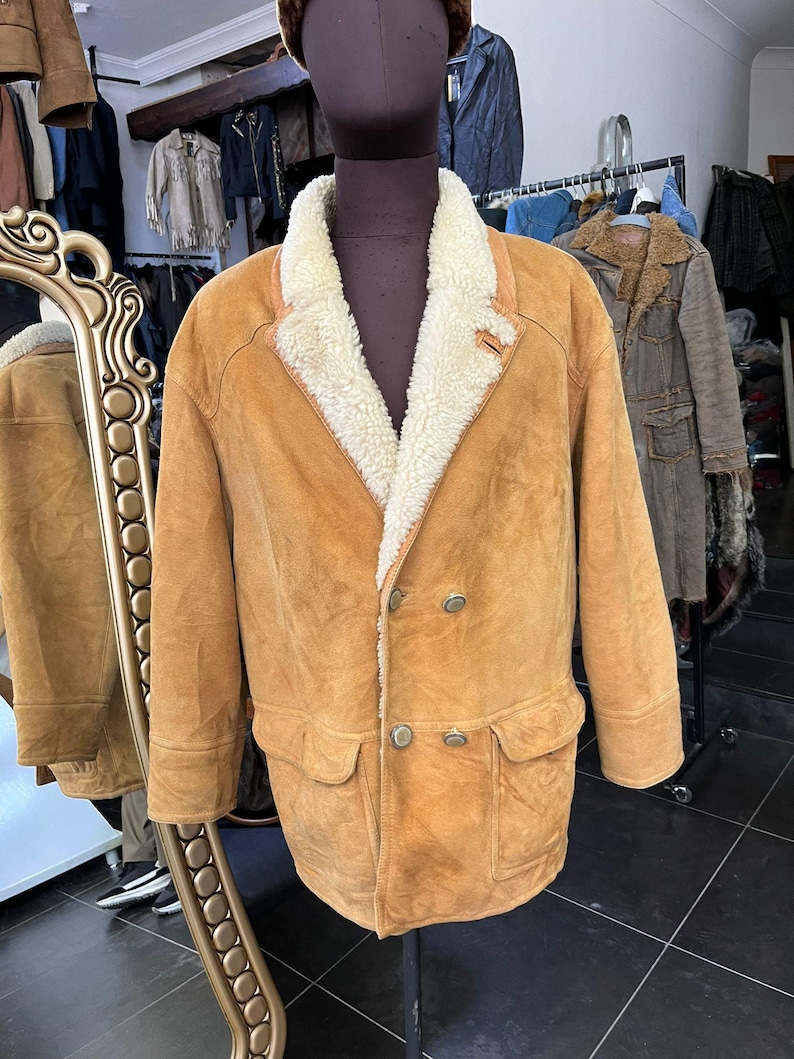 May include: A tan suede jacket with a cream shearling collar and lining. The double-breasted jacket has two front pockets and three buttons. The jacket is displayed on a mannequin in a vintage clothing shop.