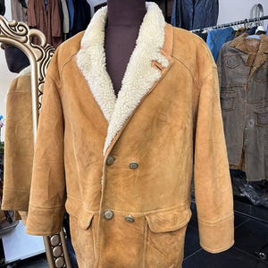 May include: A tan suede jacket with a cream shearling collar and lining. The double-breasted coat has two front pockets and large buttons. The jacket is displayed on a mannequin.