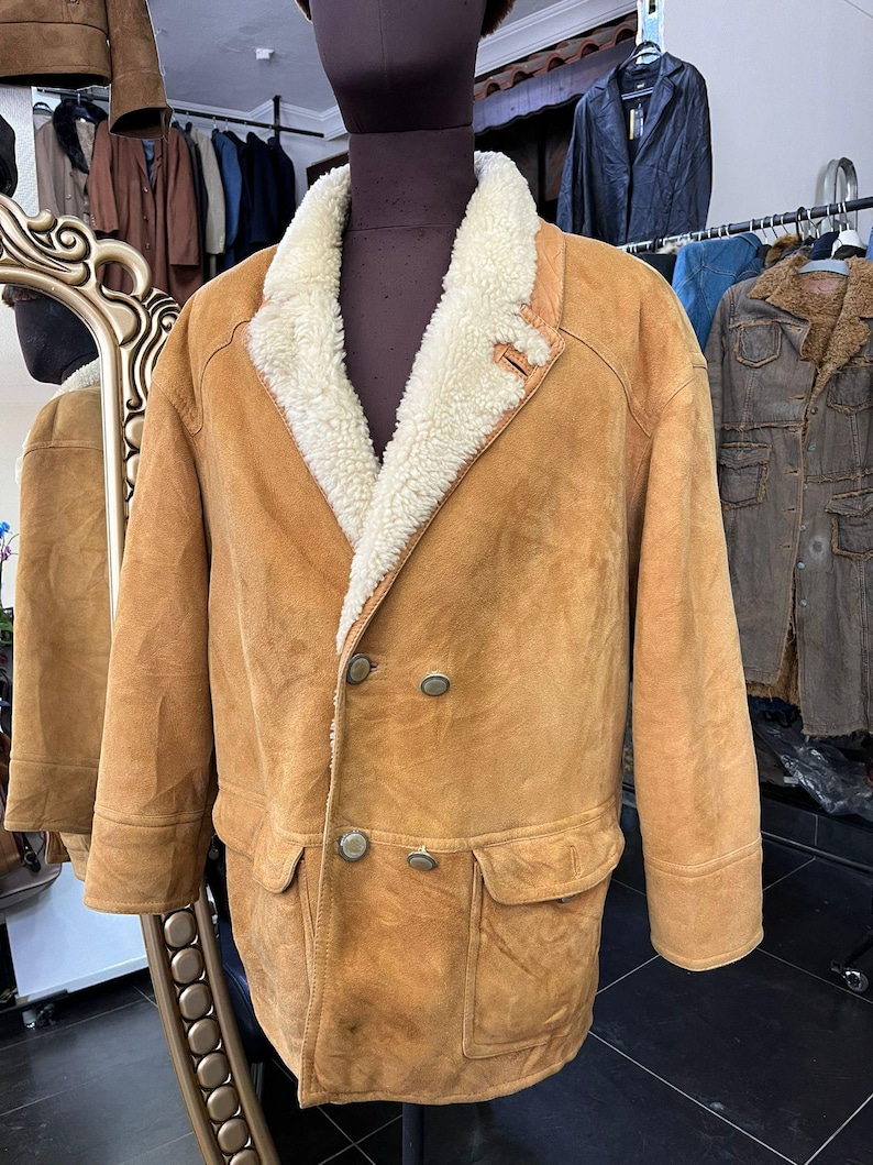 May include: A tan suede jacket with a cream shearling collar and lining. The double-breasted coat has two front pockets and large buttons. The jacket is displayed on a mannequin.