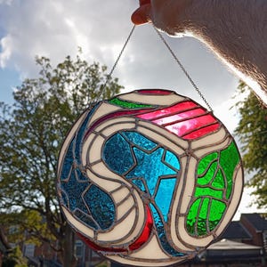 May include: A circular stained glass suncatcher featuring a geometric design with segments of textured red, blue, and green glass separated by white glass pieces. The entire piece is held together by dark metal solder lines and hangs from a thin chain.